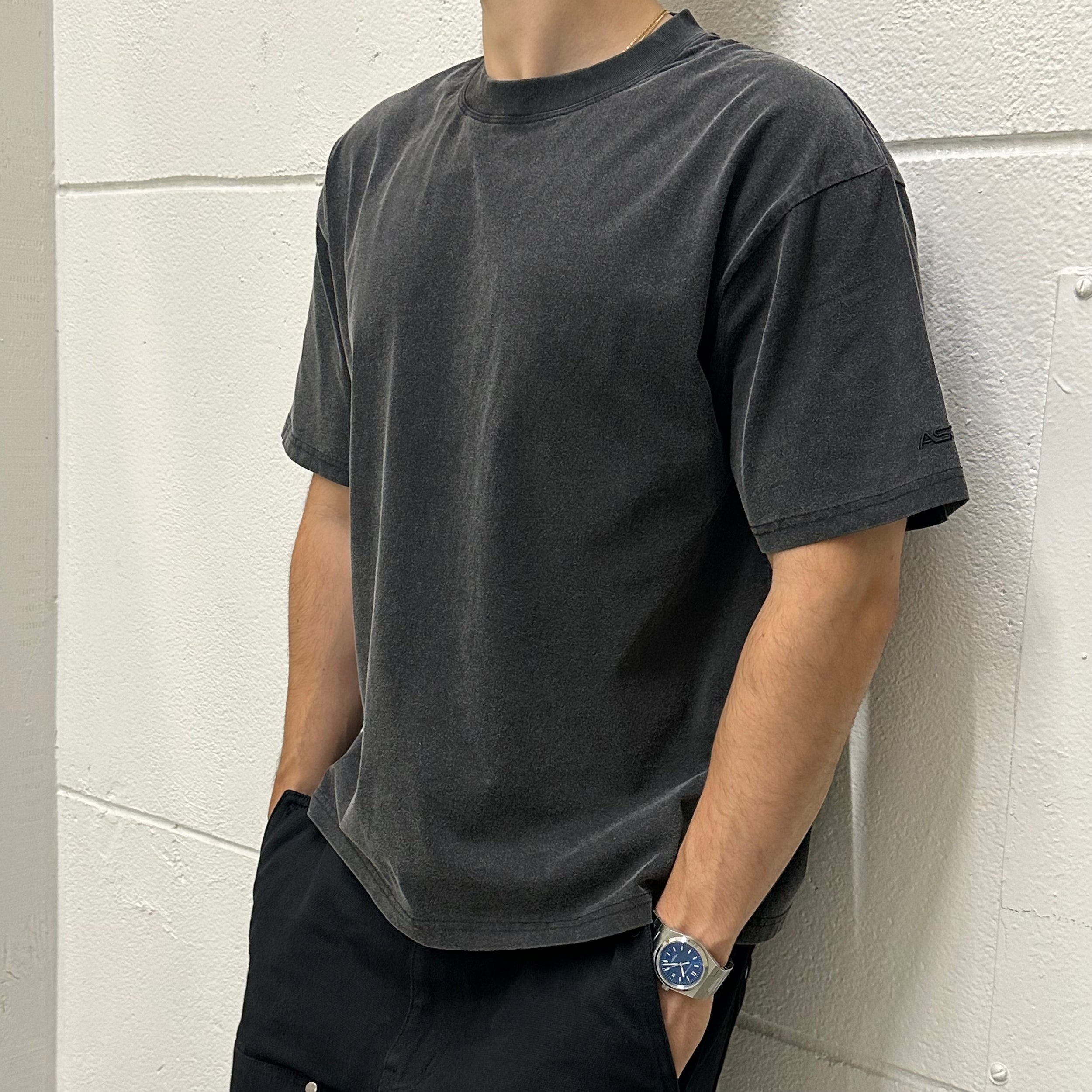 Person wearing a dark gray t-shirt and black pants against a light gray wall.